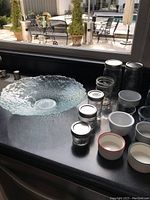 Miscellaneous kitchen glassware displayed on a countertop including clear glass canning jars with lids, large textured serving bowl, white ceramic ramekins, small dipping bowls, and a tart pan.