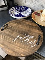 Wooden round charcuterie board with handles and 'gather' script on surface, next to the blue and white porcelain chip and dip bowl and a ceramic spaghetti bowl with cheese shaker.