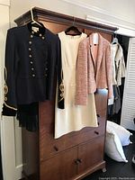 Four women's designer garments hanging on a wooden armoire: navy evening bolero-style jacket, cream wool crepe dress, pink blazer, and off-white jacket in the background.