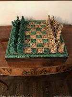 Full view of closed chess board with hand carved Mayan/Aztec themed chess pieces set up in starting positions, showing green and beige color sets, board with detailed engraved symbols, placed on wooden storage box.