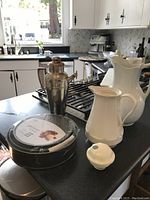 View of all lot items on kitchen counter including pewter/copper coffee server, porcelain pitchers, white thermos, springform pans in packaging, and small white lid
