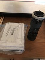 Nikon zoom lens with Nikon manual in sealed plastic bag, drum sticks visible