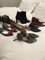 Eight pairs of assorted women's designer shoes and booties arranged on white bedding: includes black cutout ankle boots, leopard flats, black floral kitten heels, burgundy pointed toe boots, silver heels, and other styles.