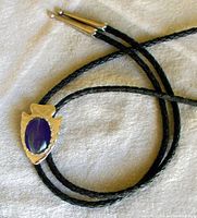 Front view of bolo tie slide with blue star sapphire-like stone and etched nickel-silver arrowhead shape, black braided leather cord visible.