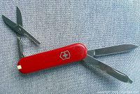 Red folding pocket tool showing scissors, file, and blade extended against textured gray background