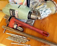 Vintage green Rockwell electric drill, red pipe wrench, silver drill bits, and a large steel railroad spike on wooden surface.