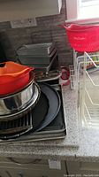 Stacked assortment of baking pans, pizza pans, racks, and silicone strainers on kitchen counter near window.