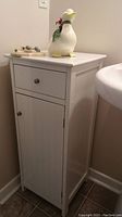 White painted wooden cabinet with upper drawer and lower door storage in corner against wall and floor tiles.