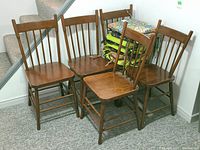 Four matching wooden dining chairs with spindle backs and one smaller chair of the same style, medium brown finish, arranged next to staircase with a stack of assorted chair pads on top.