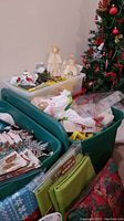 Full view of plastic totes filled with assorted Christmas decorations including angel figurines, ornaments, and artificial tree branches alongside the decorated artificial Christmas tree.