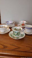 Five matched tea cups and saucers with various floral designs and gold trim on wooden surface