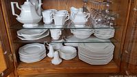 Photo showing stacked white ironstone plates of varying sizes on shelves with some bowls, cups, and saucers alongside clear stemmed glassware on upper shelf.
