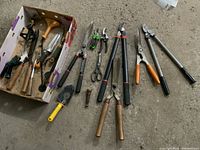 Photo showing an assortment of gardening tools on the ground including pruners, clippers, Fiskars root saw, and spray heads arranged inside and outside a cardboard box.