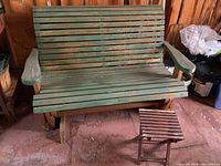 Full view of green wood slat glider bench with small matching side table beside it on rustic floor