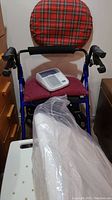 Omron blood pressure machine placed on walker seat with plaid backrest and basket visible; foam donut cushion and cover in plastic bag at front of walker.