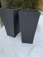 Two tall black plastic wicker planters on snow-covered ground, showing square tops and tapered bodies.