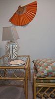 Photo displaying entire set including the rattan end table with glass top, table lamp on top, a footstool with cushion next to table, and wood and paper wall fan mounted on the wall above.