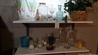 Two-tier shelving unit showing glass vases, jars, candle holders, artificial plant in woven basket, acrylic glasses, and soap dispenser.