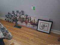 Three pieces of wall art leaning against a wall on a hardwood floor: metal leaf sculpture on the left, cityscape print in the center, framed flower print on the right.