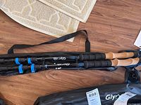 Close-up of dual Glymnis trekking poles with cork handles and blue locking mechanisms laid on a wood floor next to carrying bag.