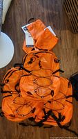 Photo showing 3 stacked orange HawLander backpacks with elastic bungee cords on the front, lying on a wood floor along with matching orange stuff sacks.