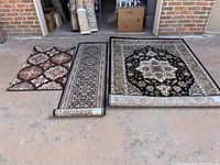 Photo showing three rugs side by side on concrete with garage background: a large 5x7 ft rug, a 2x7 ft runner, and a smaller rug with geometric pattern.