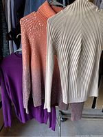 Photo of three lightweight sweaters on hangers showing cream ribbed turtleneck, rust open-knit and purple knit piece.
