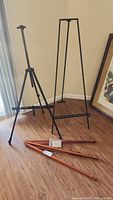 Full view of three folding easels: one black metal easel with adjustable telescoping height, one minimalist black metal easel, and three wooden easel parts on the floor.