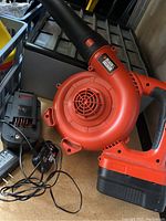 Photo showing a red and black Black & Decker cordless leaf blower with battery charger and power cord.