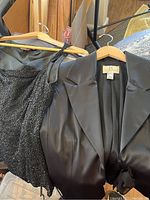 Close-up image of a black sleeveless evening dress with beaded detailing on straps and bodice, alongside a black satin blazer jacket on hangers.