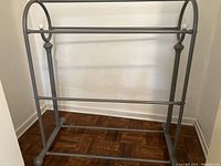 Wide shot showing full grey metal blanket rack with three horizontal bars and decorative white spherical caps at the top ends, standing on parquet flooring.