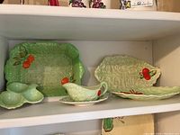 Various Carlton Ware ceramic serving plates and dishes with green leaf pattern and red fruit accents displayed on white shelves.