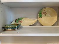 Photograph shows seven Carlton Ware ceramic pieces arranged on a white shelf: two larger serving plates with leaf motifs and a stack of five small square trinket dishes in shades of blue and green.