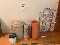 Five items on wooden surface: metal wine rack, clay wine chiller, pottery vase, small jar with wood lid, and crock with lid. Visible chips on crock.