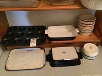 Photo showing 2 Denby baking dishes, 12 cup muffin tin, and enamel tray on wooden shelf with ceramic bowls to side.