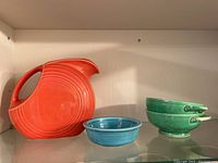 Orange Fiesta Ware pitcher, small blue bowl, and two stacked green bowls showing damage.