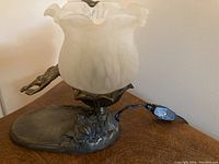 Full view of the metal lamp showing the white ruffled marbled glass shade and pond scene metal base with frog figure, electrical cord visible.