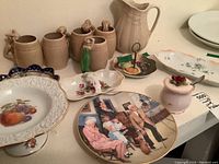 Five beige ceramic mugs with vine-shaped handles, ceramic pitcher, lidded sugar bowl, Norman Rockwell collector plate, pedestal cake stand, and various serving dishes including a small pale pink condiment pot with lid and spoon.