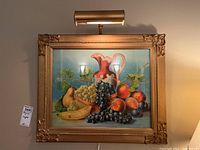 Framed print showing fruit arrangement with an overhead light attached to frame. Printed still life features grapes, bananas, pears, and peaches with a decorative pitcher.