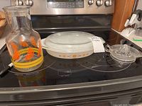 Photo of three items on stovetop: Pyrex casserole dish with lid, clear glass pitcher with orange leaf decorations, and clear glass juicer.