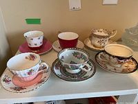 Six assorted vintage teacups and saucers arranged on white shelf