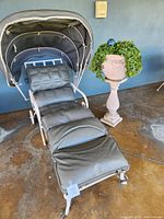 Front view of wrought iron chaise lounge with vinyl cushions and matching canopy shade alongside one pedestal planter with ivy.