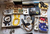 Overview of 12 small trays containing assorted necklaces, brooches, earrings arranged on a marble surface