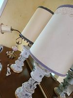Two Marbro cherub porcelain table lamps with tall cream shades trimmed in pom-poms, detailed cherub and floral bases with gold accents.