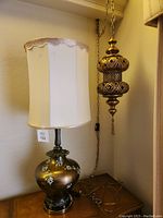 Vintage table lamp featuring brown metallic ceramic base with white and green floral details, lace-edged white lampshade, on wooden table.