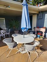 Full view of the white wrought iron round table and 4 matching chairs with a blue umbrella closed and placed in the center of the table.