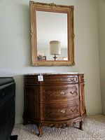 Full view of ornate gold framed mirror with mauve velvet inner frame above hand painted table with marble top and floral decorated drawers.