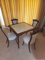Full view of the wood and white vinyl card table with four chairs arranged around it in a carpeted room with curtain backdrop.