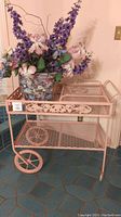 Front view of pale pink wrought iron cart with grapevine motif top rail, mesh shelves and large wheels holding the ceramic planter with faux flowers