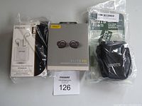Photo showing three boxed and packaged electronic items: Jabra Elite 85t earbuds in center, Roots camera bag on the right, and Escape wireless earphones on the left.